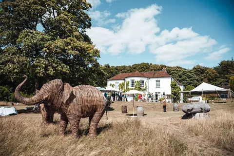 A white country house behind a meadow with a large wicker elephant sculpture