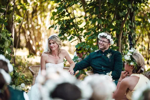 The couple seated during the ceremony under the trees, framed by guests
