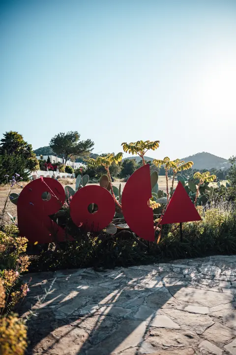 The red Roca sign among dry grass and pine trees under a blue sky