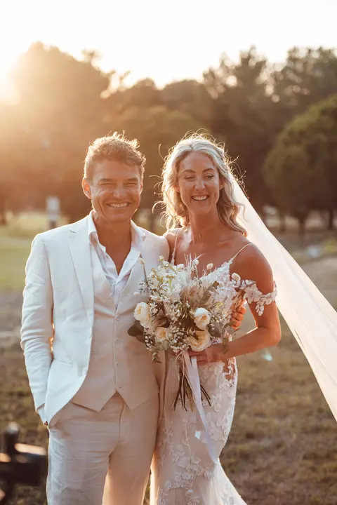 The couple standing in a field with her bouquet and a long veil