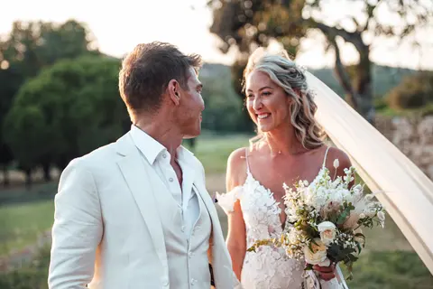 The couple smiling at each other in a field, her bouquet in hand