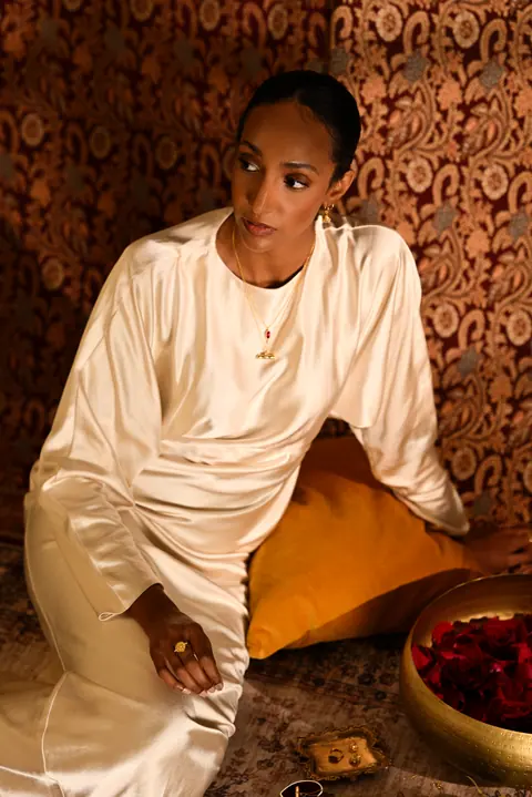 Model in ivory silk seated on a patterned carpet beside a brass bowl of fruit