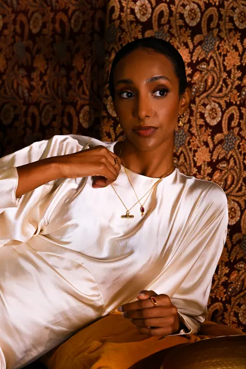 Model reclining in ivory silk against a patterned wall hanging, wearing a gold pendant