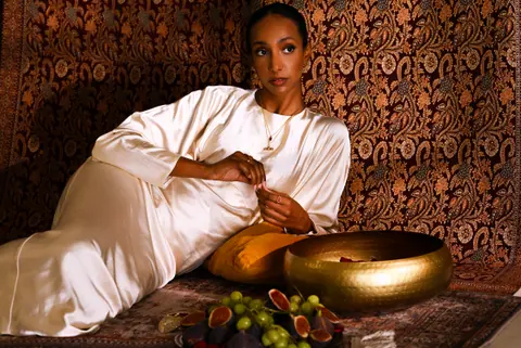 Model lounging on cushions with a brass bowl, figs and grapes in front of her