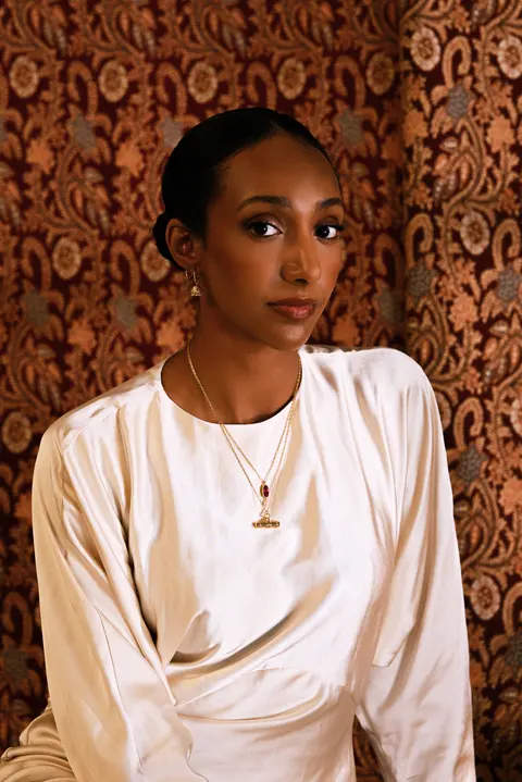 Model portrait seated on the carpet, a pendant necklace visible