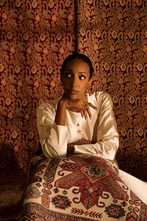 Model seated against patterned cushions, hand at her chin