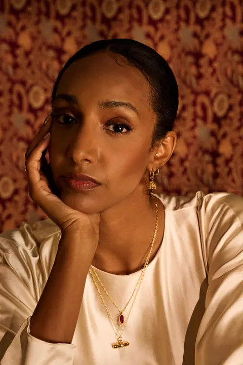Portrait of the model with her chin resting on her hand, wearing gold earrings and a pendant
