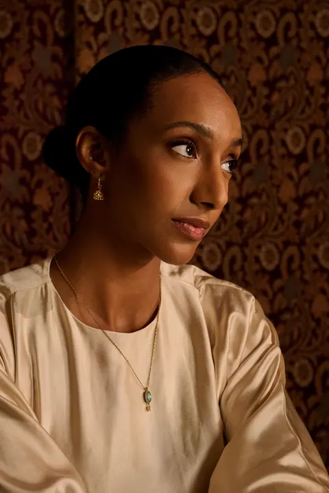Model in profile against a patterned wall hanging, a pearl drop at her collarbone