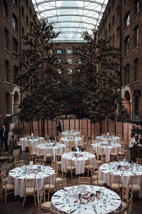 Round tables laid under a glass roofed atrium between brick walls