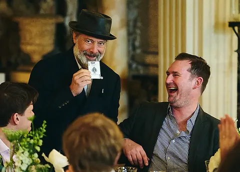 A magician in a hat showing a card to a laughing guest at the table