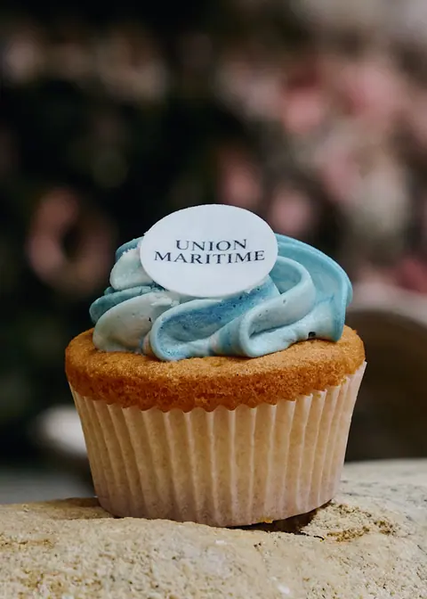 A single blue iced cupcake with a Union Maritime topper on a stone wall